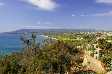 Naklejka premium A scenic view of a coastal town with a church tower and palm trees overlooking the turquoise sea and a long sandy beach, with rolling green hills in the background under a clear blue sky.