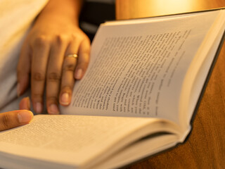 Naklejka premium Close-up of a woman's hands holding an open book with clearly visible text pages, shot in warm cozy indoor lighting with a soft glowing table lamp in the background, evoking reading and knowledge. 
