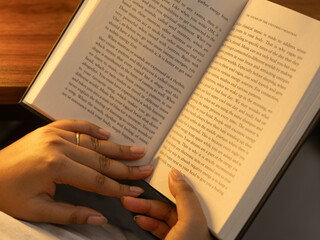 Fototapeta premium Close-up of a woman's hands holding an open book with clearly visible text pages, shot in warm cozy indoor lighting with a soft glowing table lamp in the background, evoking reading and knowledge. 