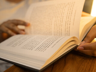 Naklejka premium Close-up of a woman's hands holding an open book with clearly visible text pages, shot in warm cozy indoor lighting with a soft glowing table lamp in the background, evoking reading and knowledge. 