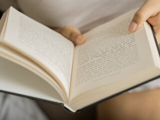 Fototapeta premium Close-up of a woman's hands holding an open book with clearly visible text pages, shot in warm cozy indoor lighting with a soft glowing table lamp in the background, evoking reading and knowledge. 
