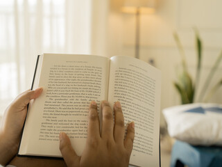 Naklejka premium Close-up of a woman's hands holding an open book with clearly visible text pages, shot in warm cozy indoor lighting with a soft glowing table lamp in the background, evoking reading and knowledge. 
