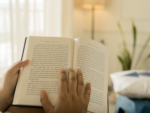 Close-up of a woman's hands holding an open book with clearly visible text pages, shot in warm cozy indoor lighting with a soft glowing table lamp in the background, evoking reading and knowledge.
