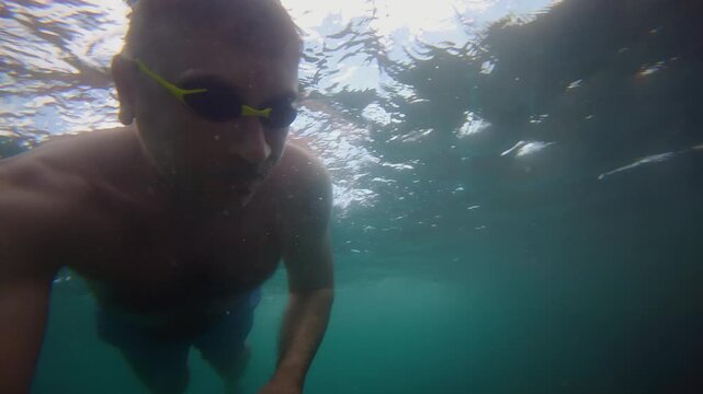 Underwater self portrait footage of a man swimming in clear blue ocean water while wearing swimming goggles