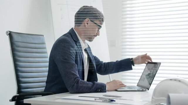 Architect or engineer working on construction blueprints and plans at an office desk, using a laptop for design and planning, embodying professionalism and project management