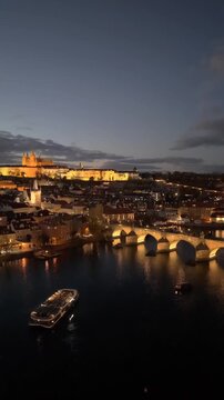 Aerial views capture the beauty of Prague as the city lights up at dusk. Iconic landmarks like Prague Castle and Charles Bridge glow against the darkening sky along the Vltava River.