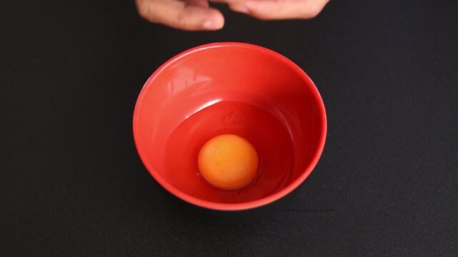 closeup human hands cracking raw egg into red plastic bowl on black kitchen counter