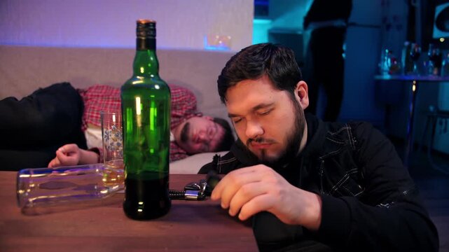 A tired drunk bearded caucasian man sits near table with a bottle of alcohol, touches his car keys, and picks up a breathalyzer. A friend is sleeping on the sofa in room. Drunk driving is prohibited