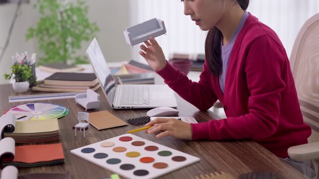 Asian women furniture designer selecting color swatches comparing paint samples working with laptop creative design professional studio workspace