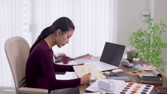 Asian furniture designer working on creative project with color palette samples laptop at modern home office workspace studio drawing sketches planning