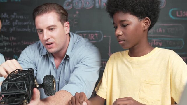 Smart teacher inspect car model construction to diverse student. high school children with mixed races listening mentor explain robotic model system at table with laptop and wires placed. Edification