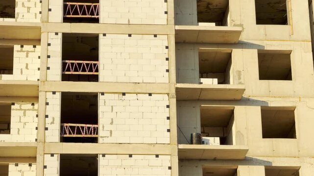 An unfinished residential concrete building stands tall under bright sunlight, capturing a raw sense of progress and anticipation. Workers lay brick walls within the empty window frames.