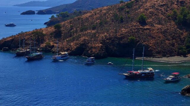 Aerial coastal view of boats resting near a rugged hillside and calm blue passage, with distant islands on the horizon, suitable for summer routes, sea travel, island tours and nature escape themes.