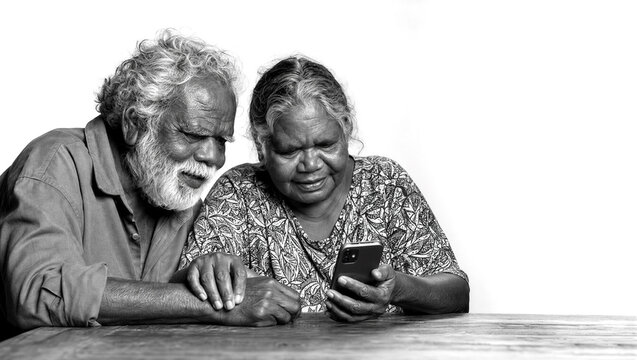 Black and white portrait of a senior Aboriginal couple looking at a smartphone screen with curious expressions while sitting at a wooden table against a white background.