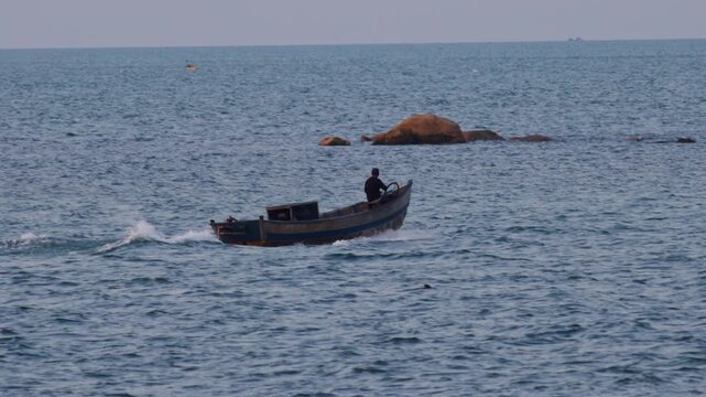 Small wooden fishing vessel with a lone fisherman at the helm sailing across the open water, creating a wake while navigating past rocky formations on the way to the fishing grounds