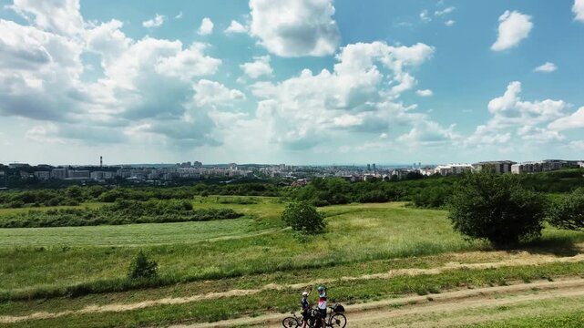 Two cyclists pause on a rural dirt road overlooking a city skyline under dramatic summer clouds. Aerial footage captures outdoor adventure and active travel