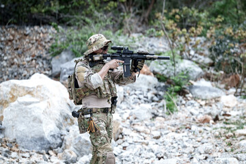 Fototapeta premium Soldier in camouflage gear aims rifle in rocky, wooded terrain