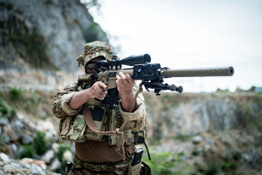 Soldier aiming a sniper rifle with scope and suppressor in a rocky outdoor environment