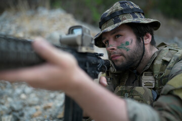 Fototapeta premium Soldier with camouflage face paint aims rifle in tactical gear during outdoor operation