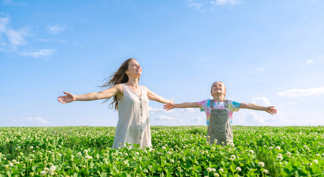 Happy mother and daughter with arms outstretched in a green clover field. Joyful family embracing nature under a blue sky