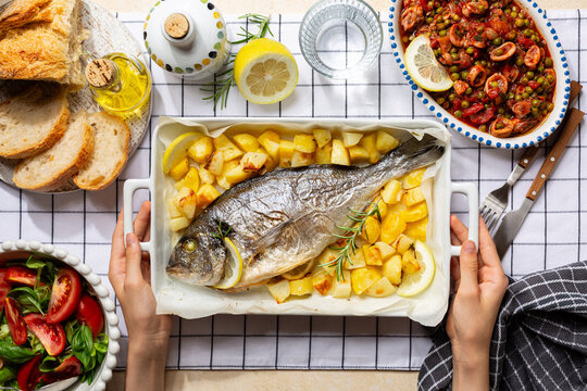 Italian, mediterranean lunch. Woman hands holding Fish traybake with sea bream or dorada fish with potato, lemon and herbs. Salad with tomato and basil, bread Squid, calamari with green Peas. 