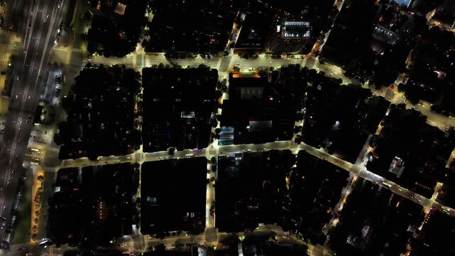 Vista cenital de una ciudad iluminada de noche con cuadr&iacute;cula de calles, manzanas, autos y tr&aacute;nsito urbano