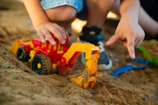 Under the evening sun, a child plays in a sandbox with a toy plastic red excavator. Close-up photo without a face, focus on an excavator toy car.