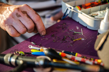 A person lays out fishing gear, mainly hooks, on a purple surface. Fishing floats, sinkers, a spinning rod, a fishing box and a tube of glue are visible. © Rystemys