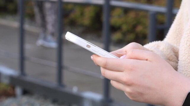 Hands hold phone, focused on screen outdoors. Background shows blurred fence and dog walking away. Person wears light-colored jacket and smartwatch. Dog moves slowly, appearing fluffy with light coat