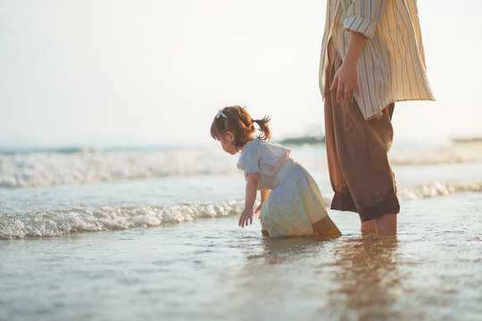 Cute little Asian girl playing with sea waves on sandy beach with her mother at sunset, happy family enjoying tropical beach vacation during golden hour, outdoor exploration, active childhood growth