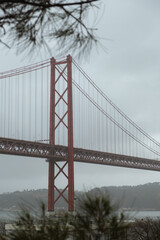 Fototapeta premium View of the 25 de Abril Bridge in Lisbon stretching across the Tagus River on a cloudy day, with soft mist surrounding the iconic red suspension structure and calm urban waterfront scenery.