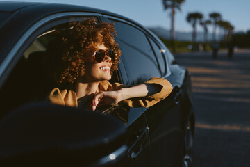 Fototapeta premium Smiling woman with curly hair leaning from a car window, enjoying a sunny drive. Retro film color mood enhances a carefree lifestyle and warm, optimistic vibe. Perfect for lifestyle scenes.
