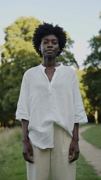Vertical video of a young Black woman with natural hair standing in a park. Portrait of a woman wearing a white linen tunic and beige trousers. Natural beauty and sustainable lifestyle concept