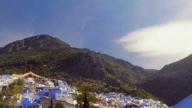 Drone rises from Chefchaouen street toward Rif Mountain peak, palm tree and blue sky with clouds