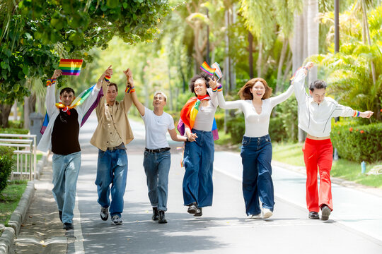 Happy diverse LGBTQ friends holding hands running and celebrating with rainbow pride flags on a sunny park pathway during summer pride month festival