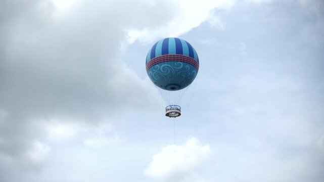Balloon floating high above the clouds carries passengers in an elevated observation basket. Upward view highlights tethered balloon ride, aerial attraction, sightseeing experience, and overcast sky
