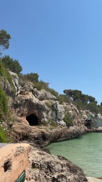 Coastal rocky cove in Cala Ferrera, Mallorca with limestone cliffs, sea cave, turquoise Mediterranean water and pine trees under clear blue sky on a sunny summer day.