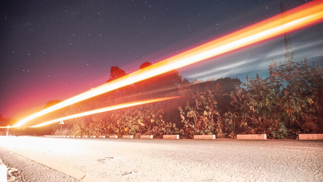 A starry night sky above a forest road, with vivid red and orange light trails streaking across, capturing motion and speed against the stillness of trees and stars. Orange light trails.