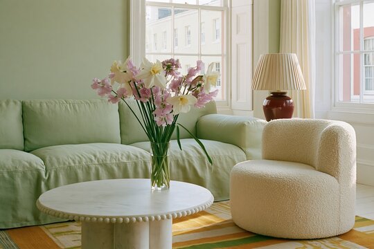 Living room featuring a soft sage linen sofa, round marble coffee table, and a textured armchair, with a vase of flowers and natural light from large windows