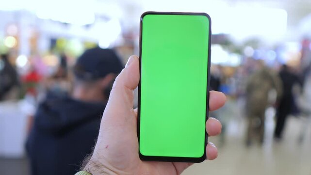 Hand holds phone with green screen. Background shows blurred crowd in public space. Phone center focus. Ideal for ads, UI mockups, or app demonstrations. People appear distracted or moving casually