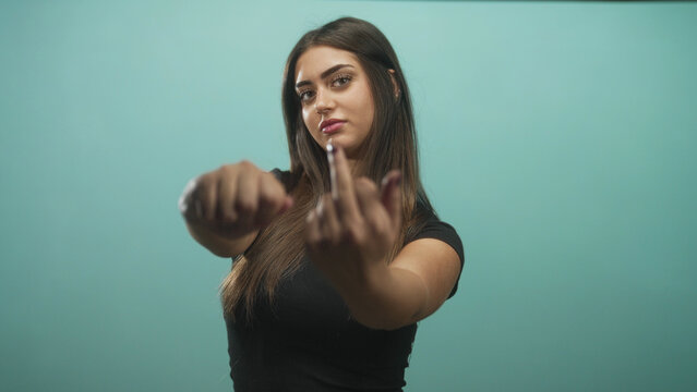Woman young hispanic with long hair extends middle finger toward camera in studio teal backdrop wearing black t shirt, direct stare; defiance attitude.