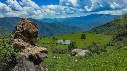 Fototapeta premium Beautiful panoramic view of a vibrant green mountain valley featuring a small clear lake and a lone tree. Textural rocks covered with lichen in the foreground. Majestic blue ridges and fluffy white cl