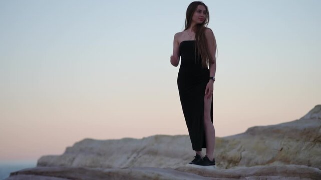 Beautiful woman with long hair in an evening dress dancing sensually on a rocky cliff against the scenic sunset sky over the sea