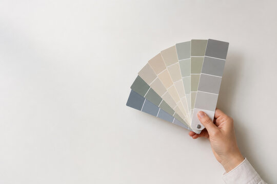 Hand holds neutral color swatches on white wall. Space for text.