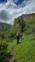 Obraz premium Back view of active tourists with backpacks hiking along a narrow dirt trail on a lush green mountain slope. Majestic rocky cliffs and high peaks rise in the background under a cloudy summer sky. Conc