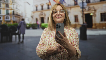 Fototapeta premium Woman holding a smartphone with hand, smiling and taking a selfie in a busy street with historic building facades behind; contentment.