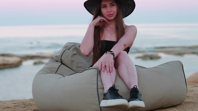 Attractive smiling woman sitting on a comfortable beanbag chair on the sandy beach, enjoying her seaside vacation during a beautiful sunset