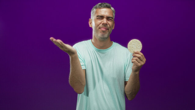 Man holding a round corn pastry in one hand and pinching his nose with the other in a purple studio set, face cringing at the smell; disgust.