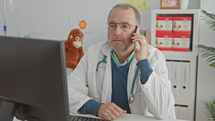 Fototapeta premium Man doctor holding smartphone to ear speaking on phone with stethoscope and computer visible in building; concerned telemedicine support.