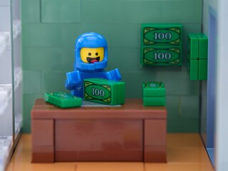 Obraz premium Tambov, Russian Federation - May 26, 2025 A blue Lego astronaut minifigure counts green toy cash bundles at a brown wooden desk.
